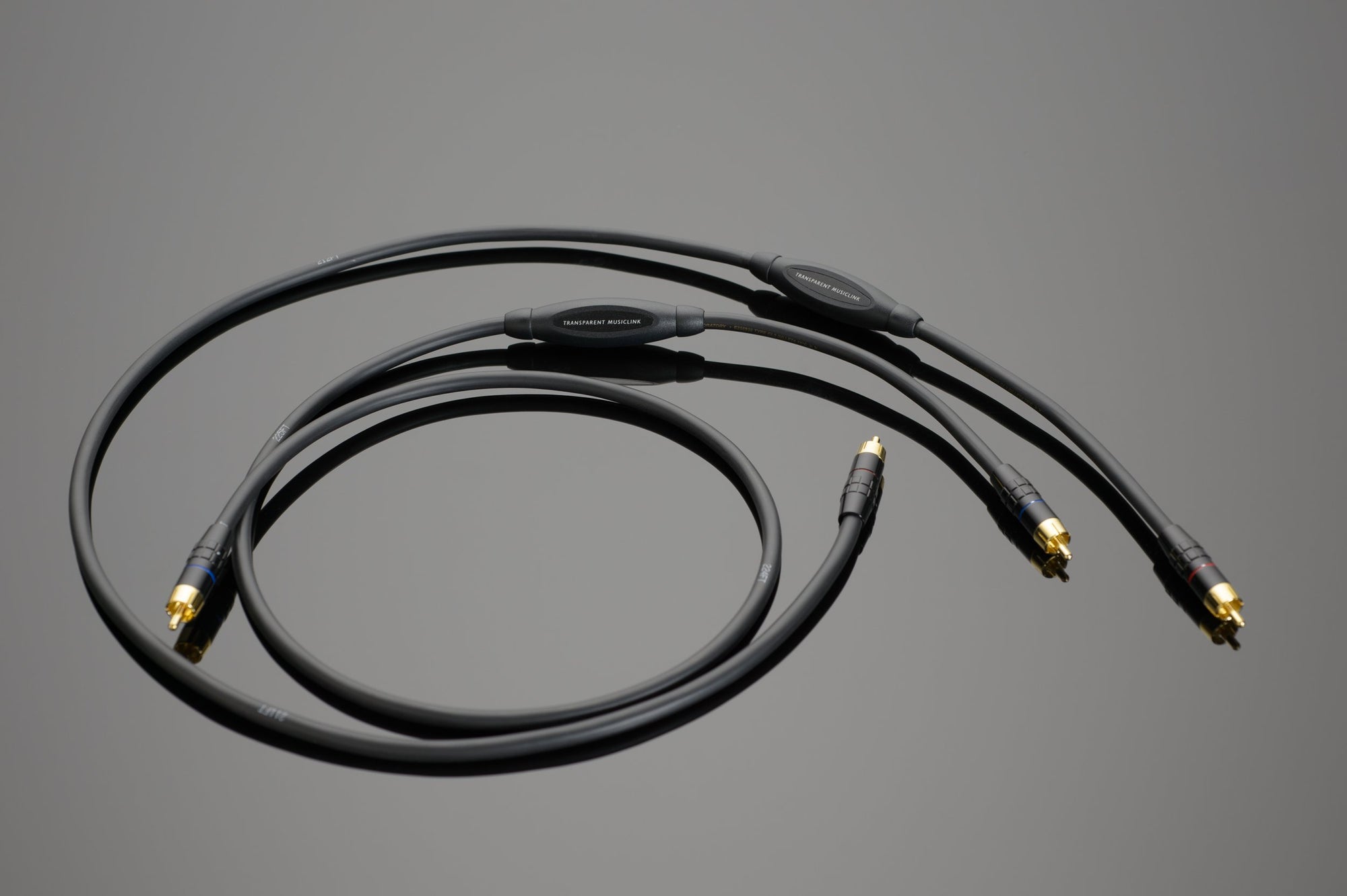 The MusicLink RCA Interconnect by Transparent Audio, a coiled black audio cable with gold-plated connectors designed to enhance your music system, lies elegantly on a gray background.